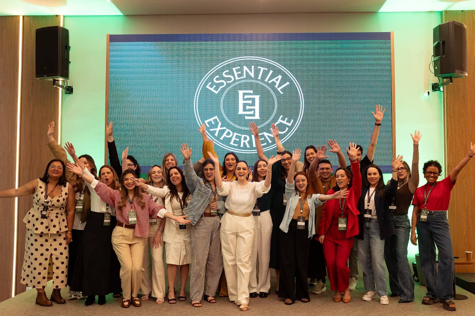 Dezenas de participantes do Essential Experience de braços para o alto em frente ao telão